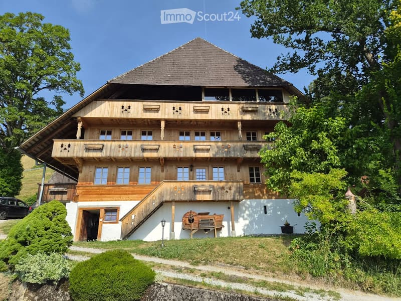 A two-story wooden house with a steep roof, multiple windows, and balconies, surrounded by greenery and trees.