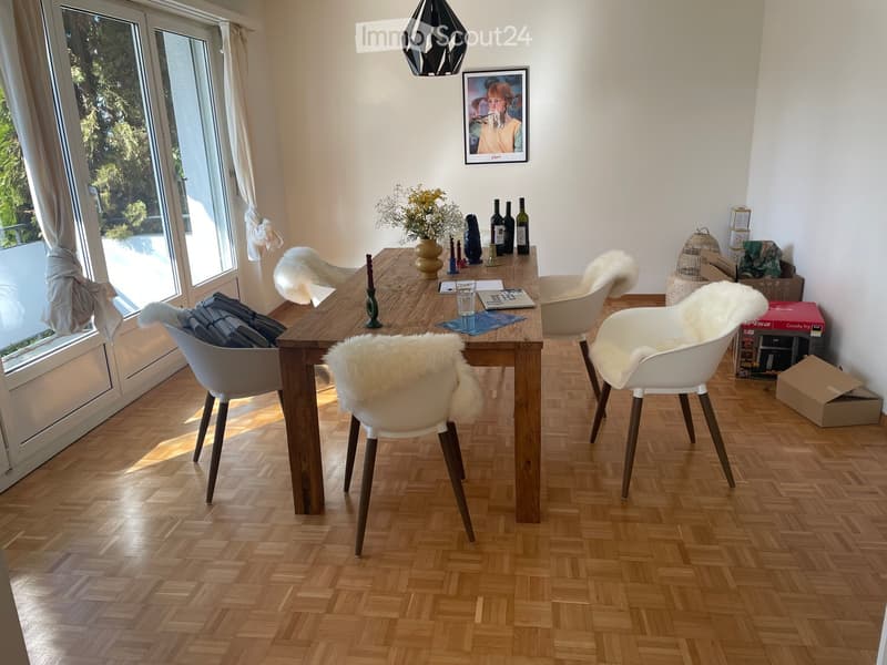 Dining room with wooden table, white chairs, parquet floor, large windows, picture frame, flowers, wine bottles, boxes
