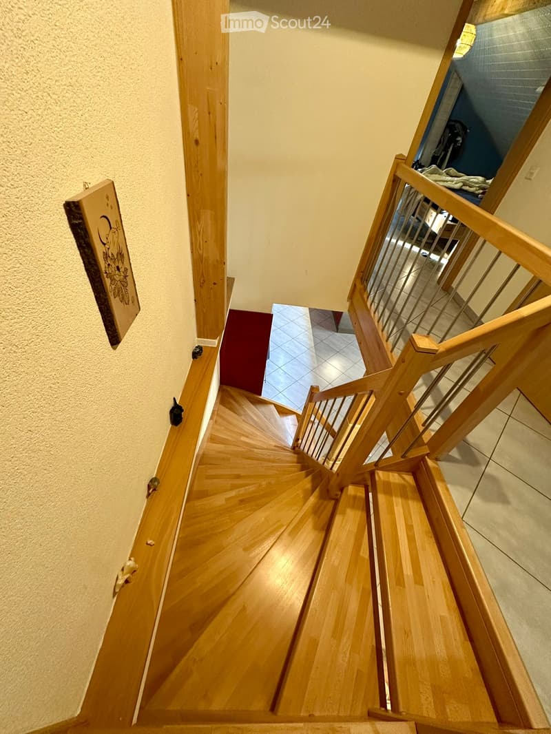 Spiral wooden staircase with metal railing, beige walls, and a red carpeted landing leading to an adjacent room.