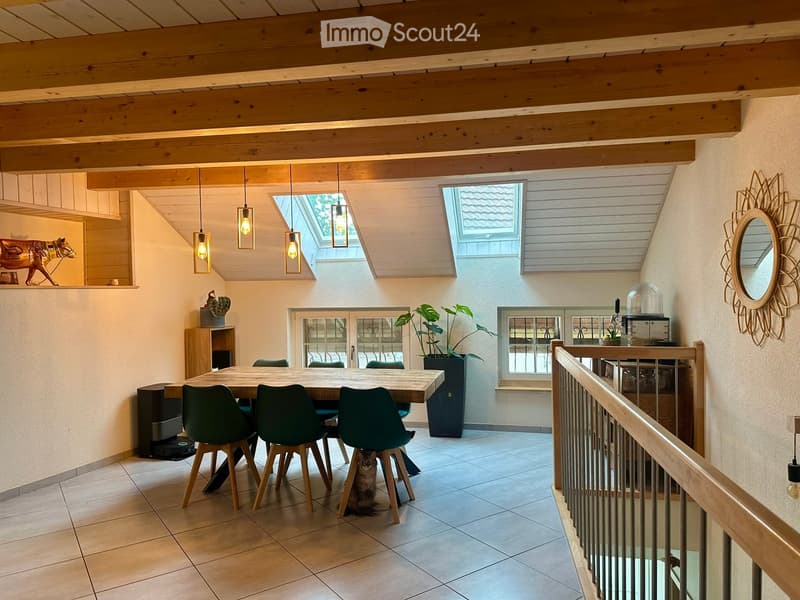Dining area with a wooden table, green chairs, pendant lights, and a cat sitting under the table. There is a staircase railing on the right and a large window with a view.