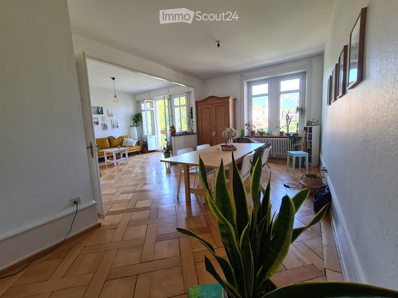 Bright living and dining area with wooden flooring, large windows, and a balcony door. Features a yellow sofa, white coffee table, dining table with chairs, and various decorative items.