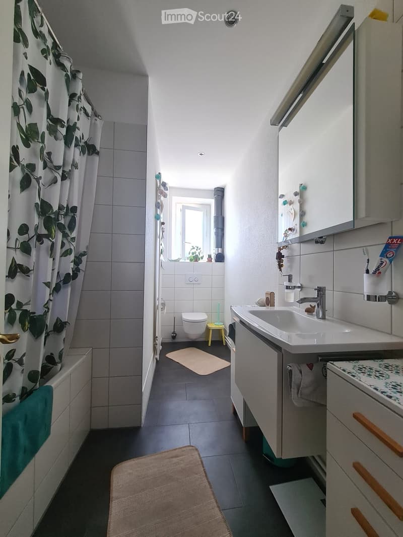 Modern bathroom with a bathtub, sink, toilet, and mirror. Features a floral shower curtain, a small window, and a bathroom rug on the floor.