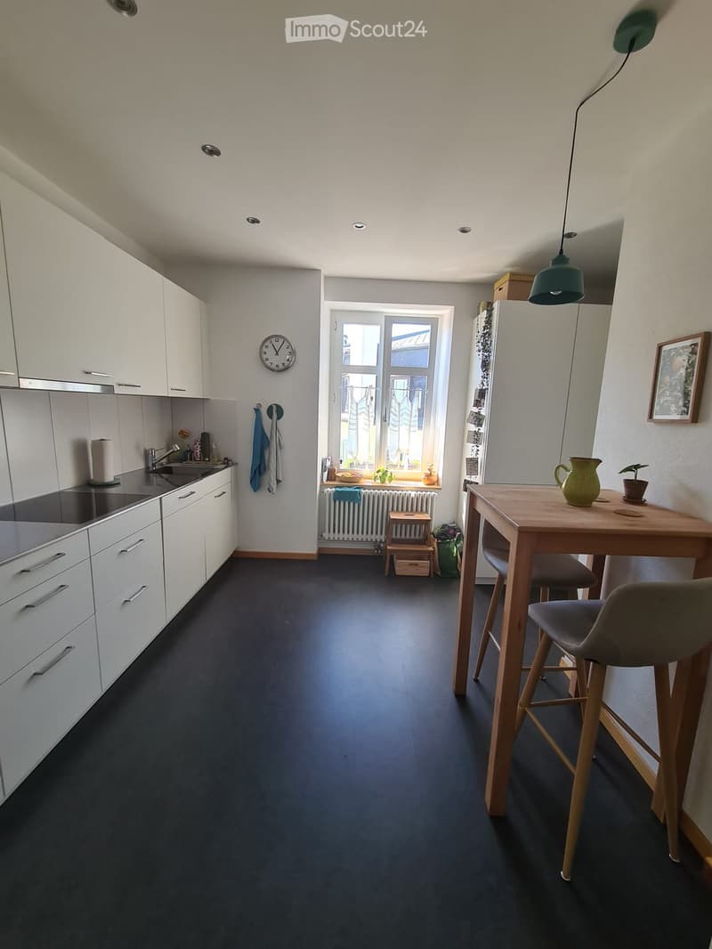 Modern kitchen with white cabinets, dark grey floor, and a small dining area with a wooden table and chairs. Features a sink, faucet, clock, and potted plant on the windowsill.