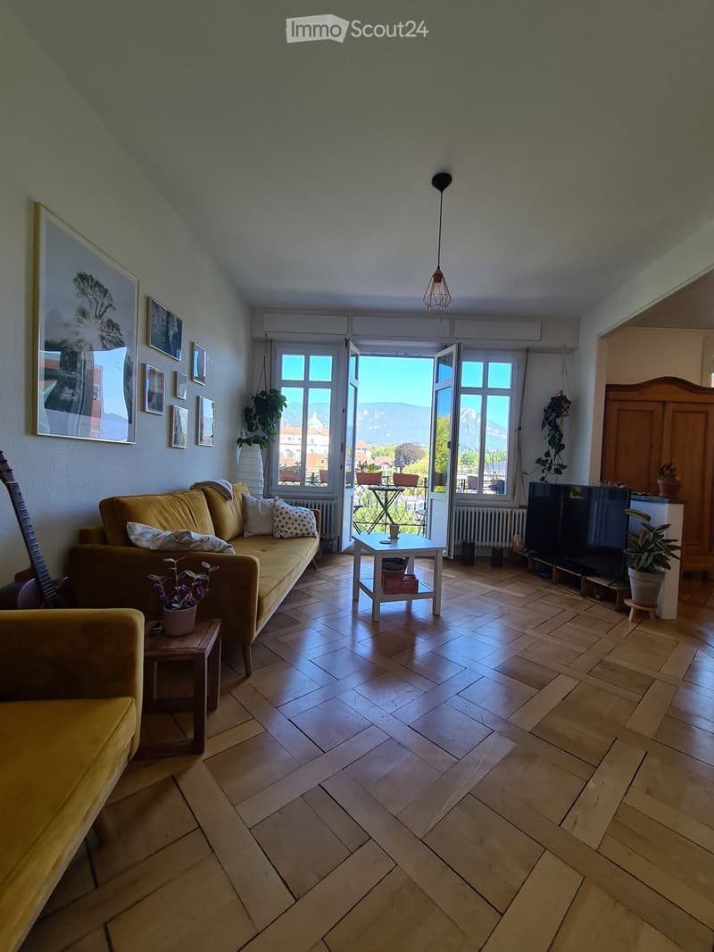 Bright living room with wooden flooring, large windows offering a scenic view, a yellow sofa, a coffee table, a guitar, a television, and several plants.