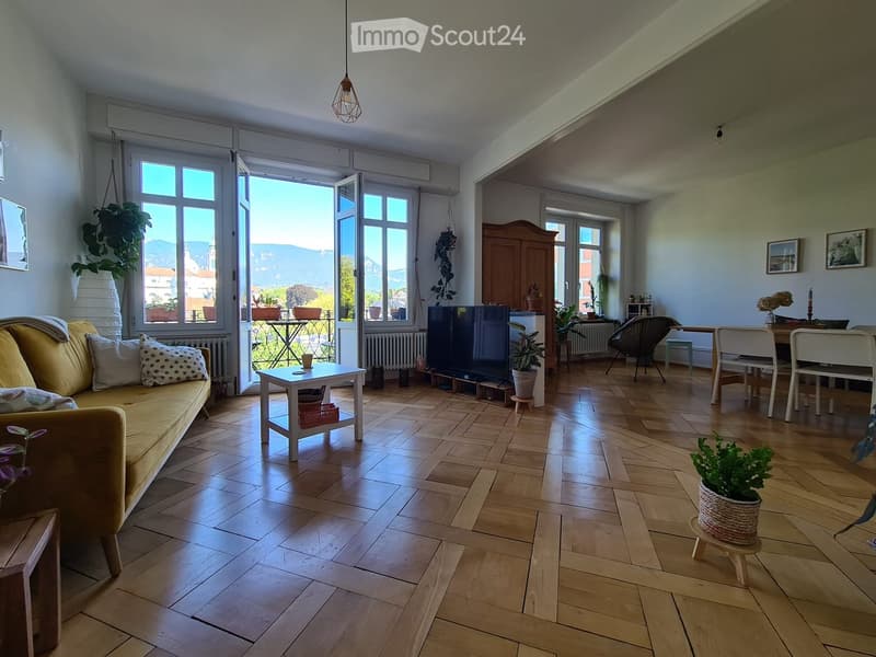 Bright living room with wooden flooring, large windows, a yellow sofa, coffee table, dining area, and potted plants.