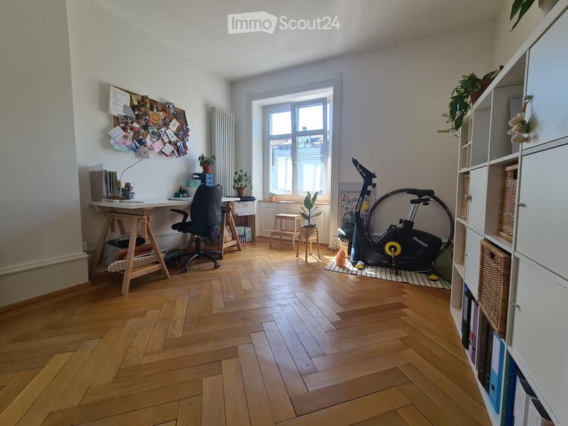 A multi-functional room with a wooden floor, a desk with a chair, an exercise bike, a shelf with books and baskets, a small stool, and a plant near the window.