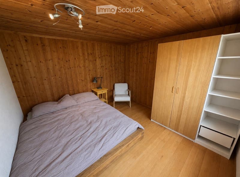 Cozy bedroom with wooden walls and ceiling, featuring a bed with pink bedding, a white armchair, a small wooden nightstand with a lamp, a built-in wardrobe, and a white shelving unit.