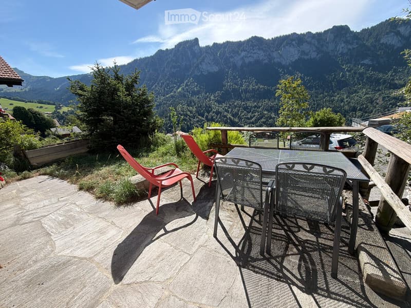 Balcony with stone floor, wooden railing, and outdoor furniture including a table and chairs, overlooking a scenic mountain landscape.