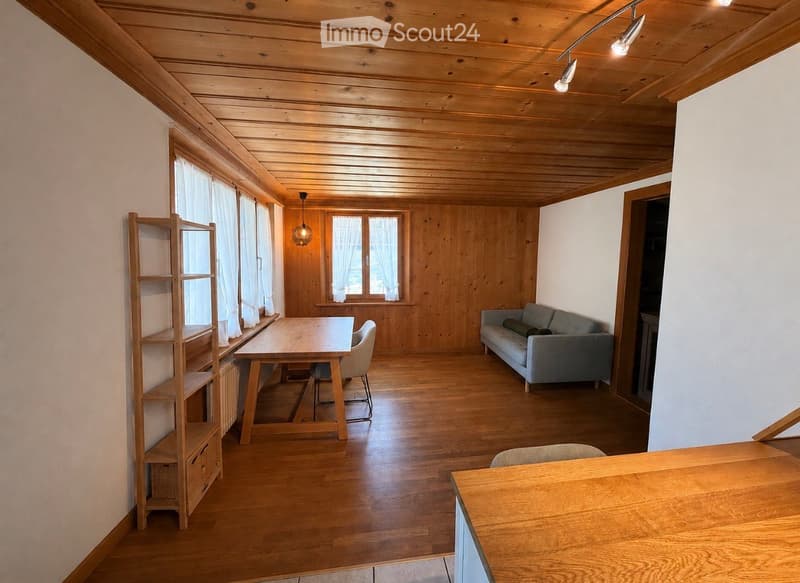 Bright living room with wooden walls and ceiling, large windows with white curtains, a grey sofa, a wooden dining table with chairs, and a wooden shelf.