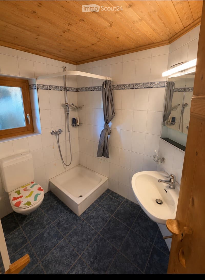 Bathroom with white tiles, a bathtub, a toilet with a colorful seat cover, a sink with a modern faucet, a shower area with a glass enclosure, and a towel hanging on the wall.