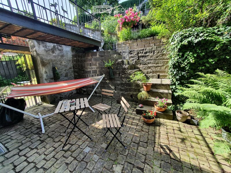 A cozy outdoor garden area with a hammock, a small table with chairs, and potted plants, surrounded by stone walls and greenery.