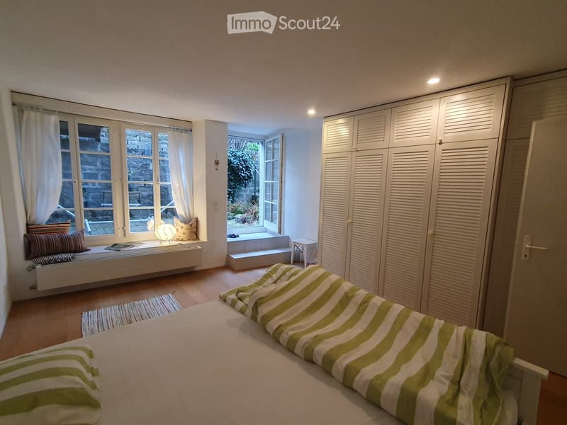 Cozy bedroom with a bed, striped duvet, white wardrobe, small table and chair by the window, and a bench seat with pillows.