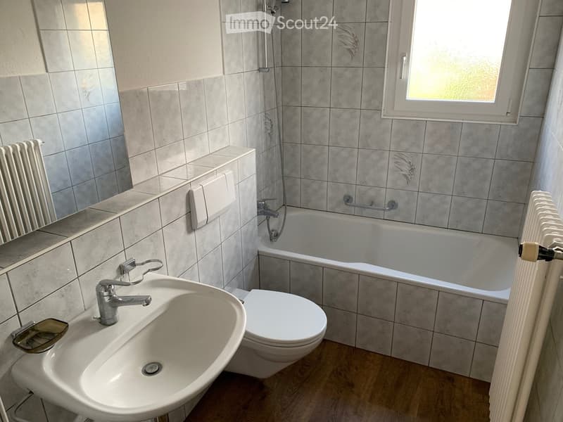 Bathroom with a sink, toilet, bathtub, radiator, and mirror. The walls are tiled in a light grey color, and the floor is wooden.