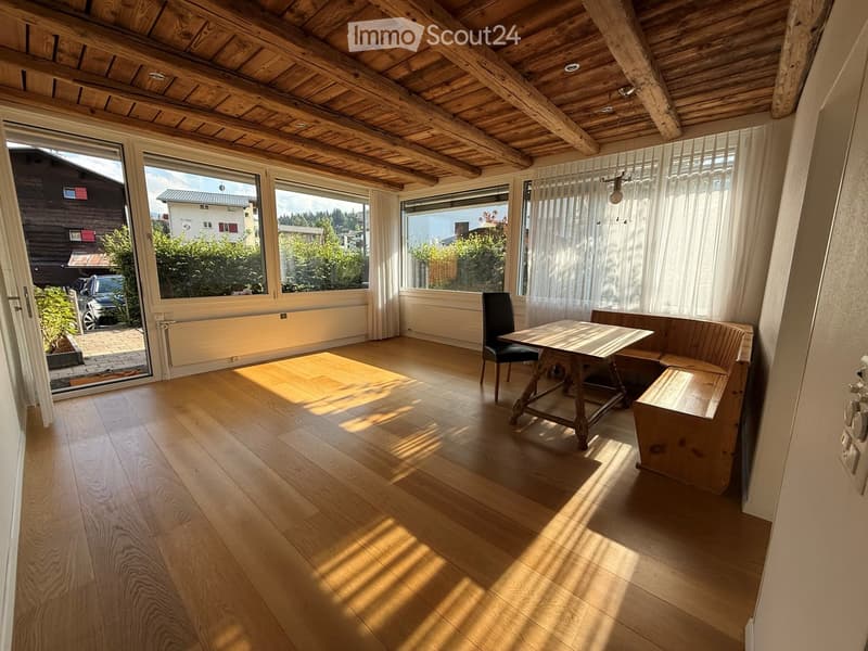 Bright room with wooden floor, large windows, a wooden table, a chair, and a bench. The room has a wooden ceiling with exposed beams and a view of the outdoors.