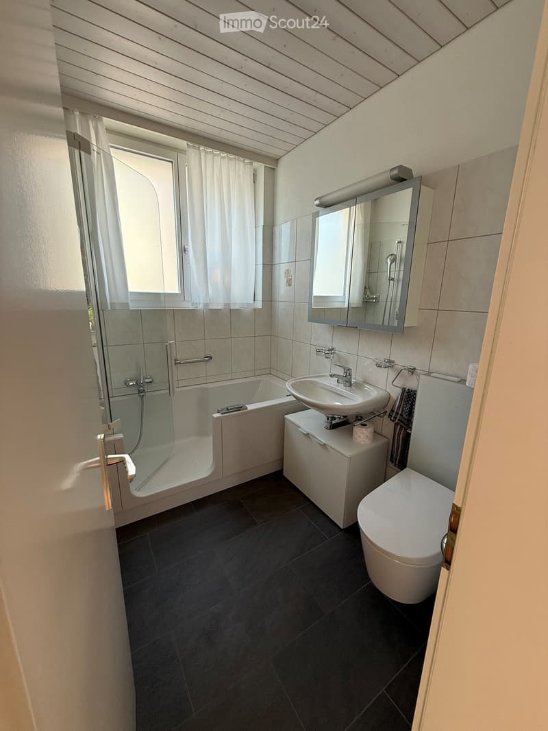 Modern bathroom with dark tiled floor, white bathtub, sink, toilet, mirrored cabinet, and a window with white curtains.