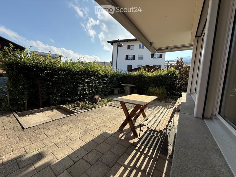 Balcony with a wooden table and bench, surrounded by greenery and a view of neighboring houses.