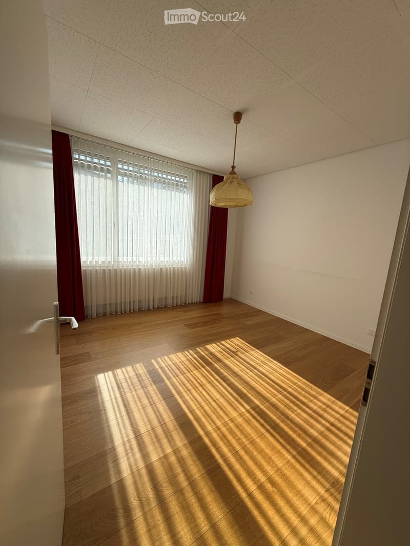 Empty room with wooden flooring, white walls, and a hanging light fixture. Sunlight streams in through large windows covered with sheer white curtains and red drapes.