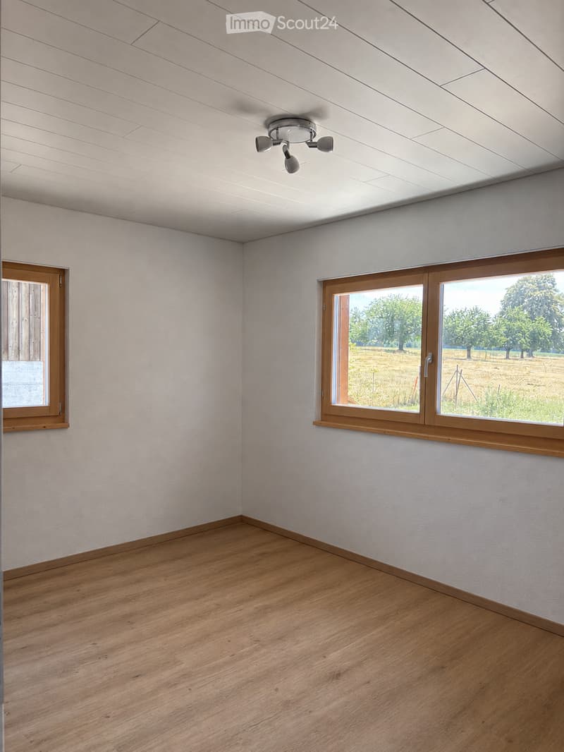 Empty room with light wooden flooring, white walls, and large windows offering a view of a green field. The ceiling features recessed lighting.