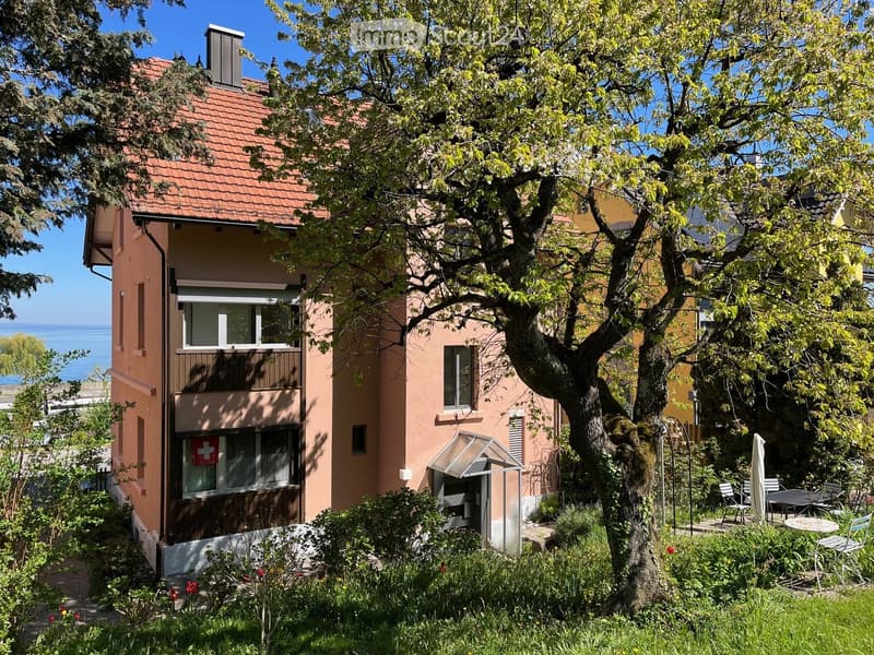 Two-story house with red-tiled roof, beige walls, and large windows. Features a balcony, garden with a large tree, outdoor seating area, and a view of the sea.