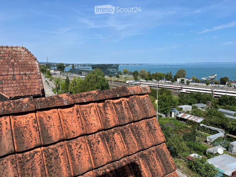 View from a rooftop terrace showing a body of water, distant buildings, train tracks, and surrounding greenery.