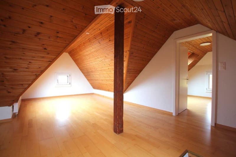 Empty attic room with wooden floors and slanted wooden ceilings, featuring exposed wooden beams and white walls.