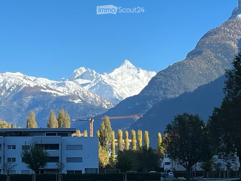 Modern multi-story building with large windows and a flat roof, surrounded by trees and set against a backdrop of snow-capped mountains.