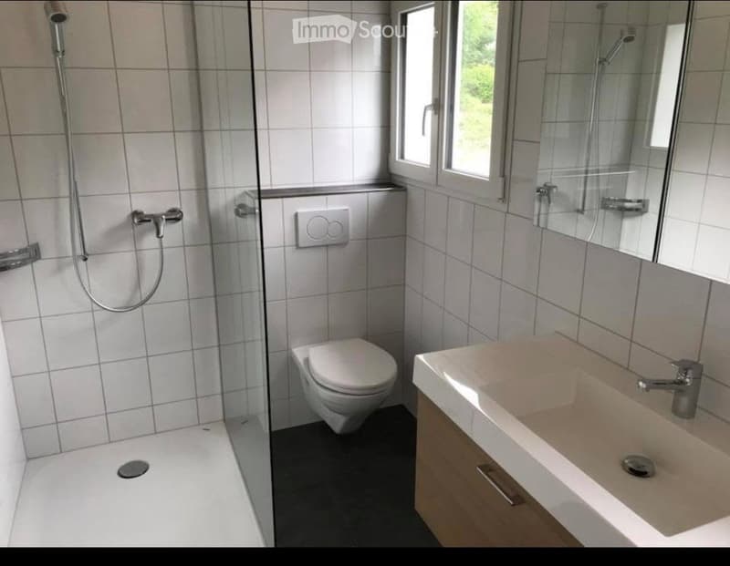 Modern bathroom with white tiles, a toilet, a sink with a mirror, and a shower area with a glass enclosure.