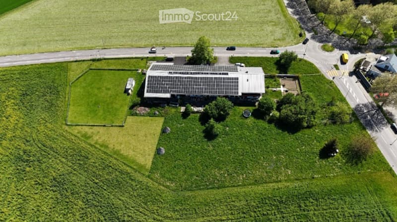 Aerial view of a modern building with a solar panel roof, surrounded by green fields and a road nearby.