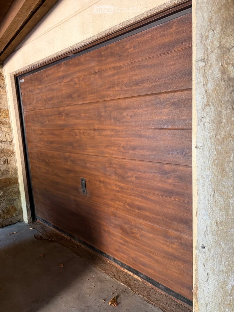 Wooden garage door with a textured finish, mounted between two stone walls, with a concrete floor visible beneath.