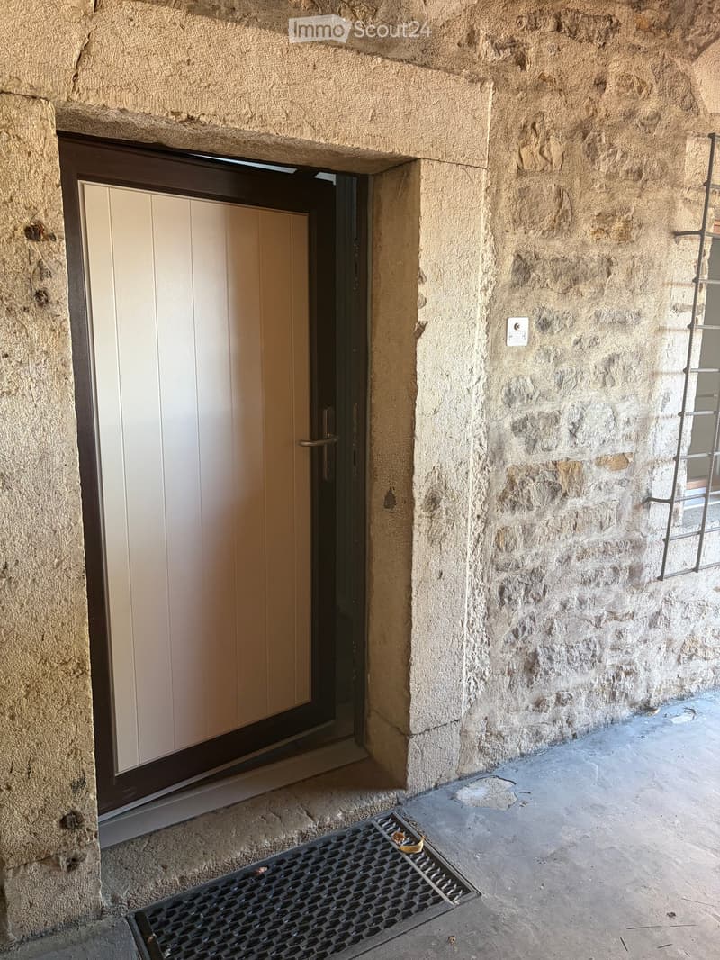 A narrow stone-walled corridor with a wooden door at one end and a metal grate on the floor.