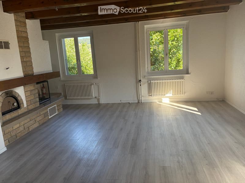 Empty room with wooden flooring, exposed wooden ceiling beams, and a brick fireplace. Two windows with white frames and radiators beneath them.