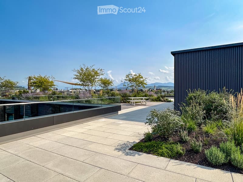 Modern rooftop garden with stone tiles, glass railing, small seating area, and lush greenery.