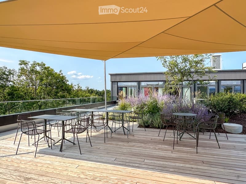 Wooden deck with tables and chairs, large orange canopy, glass railing, and lush greenery in the background.
