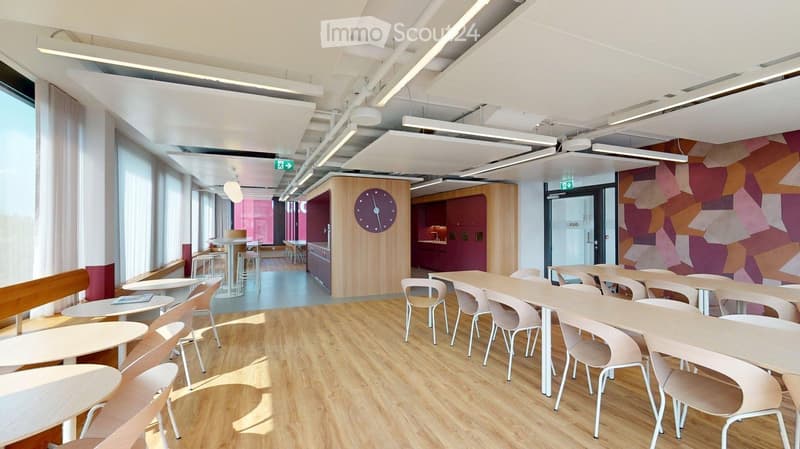 Bright, modern room with wooden flooring, white tables, and chairs arranged in rows, a clock on a wooden partition, and a colorful geometric wall design.