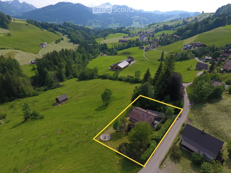 Aerial view of a rural house surrounded by green fields and trees, with a driveway and nearby structures visible.