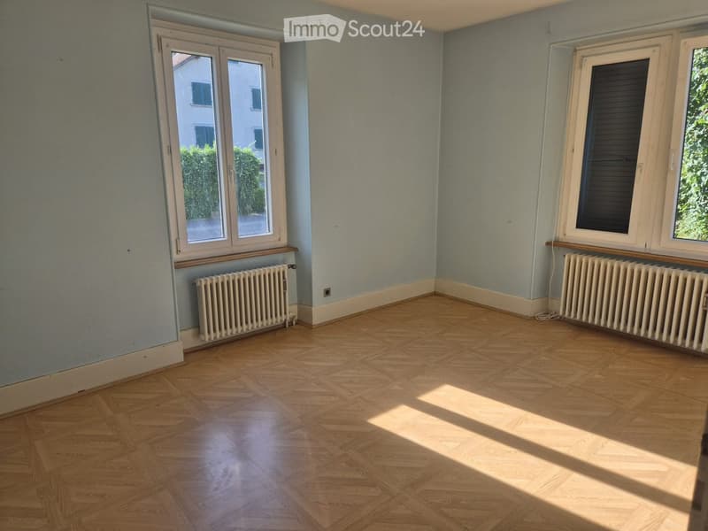 Empty room with wooden parquet flooring, light blue walls, two radiators, and large windows with shutters. The room is completely unfurnished.