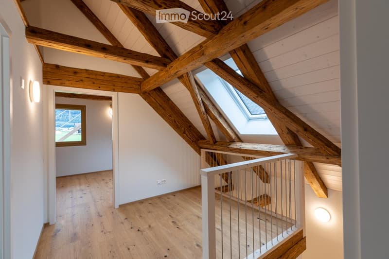 Bright, airy hallway with exposed wooden beams, wooden flooring, and a skylight window providing natural light. Features a modern metal railing and built-in lighting.