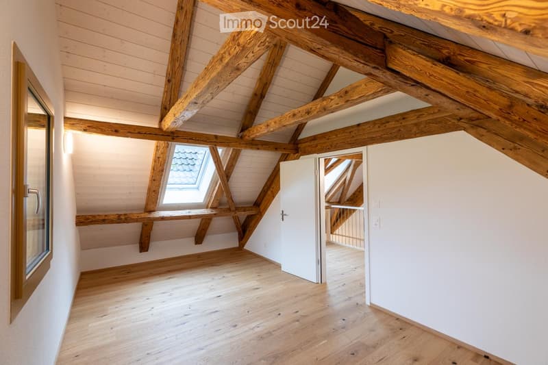 Bright, empty attic room with exposed wooden beams, wooden flooring, and large skylight windows. The space features white walls and a door leading to another area.