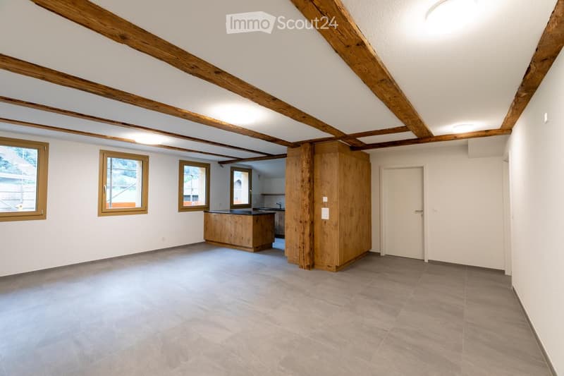Bright, open room with exposed wooden beams, tiled floor, and large windows. Includes a small kitchen area with cabinets and a sink.