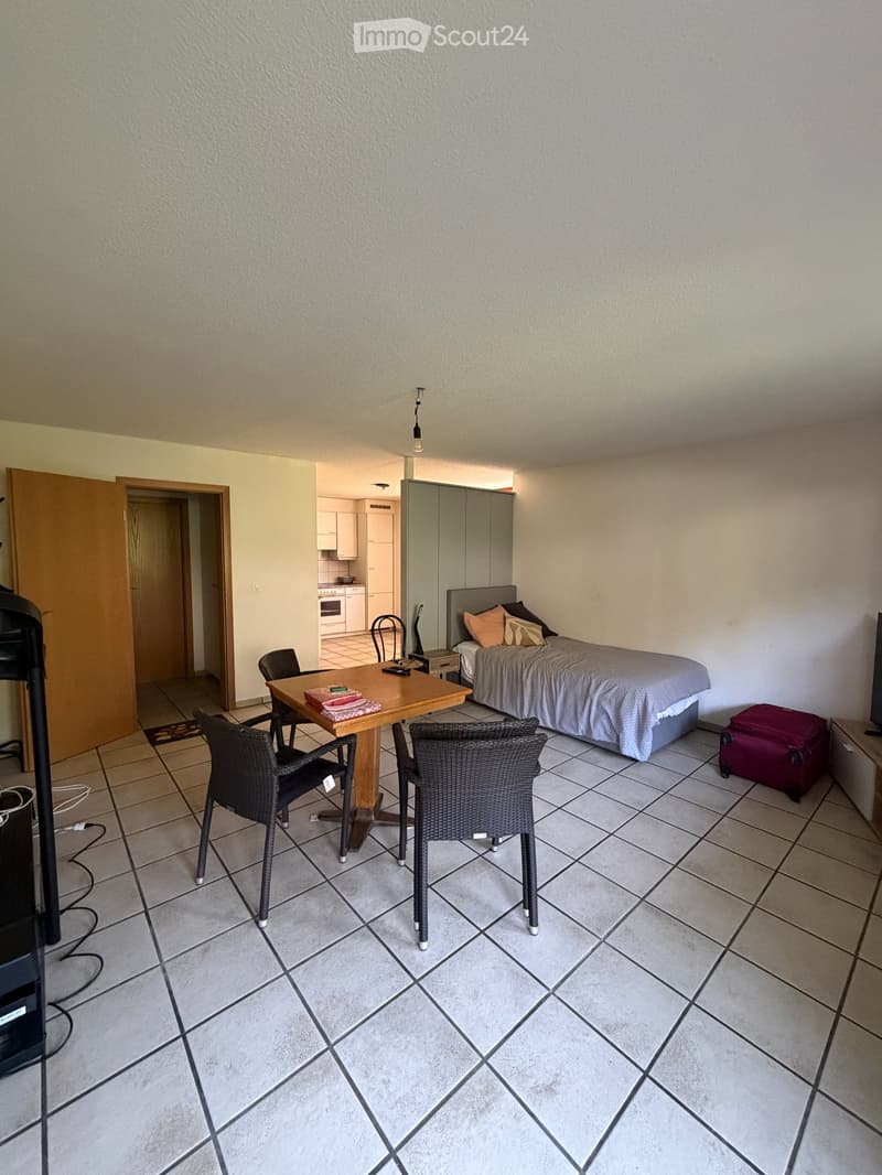A furnished room with a bed, a small table with chairs, a nightstand, and a suitcase. The room has tiled flooring and a view into an adjacent kitchen area.