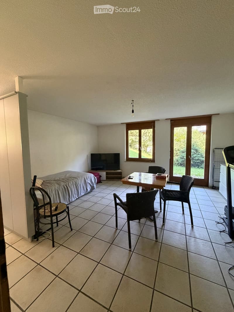 A furnished room with tiled flooring, a sofa, a table with chairs, and a television. The room has large windows and a sliding glass door leading outside.