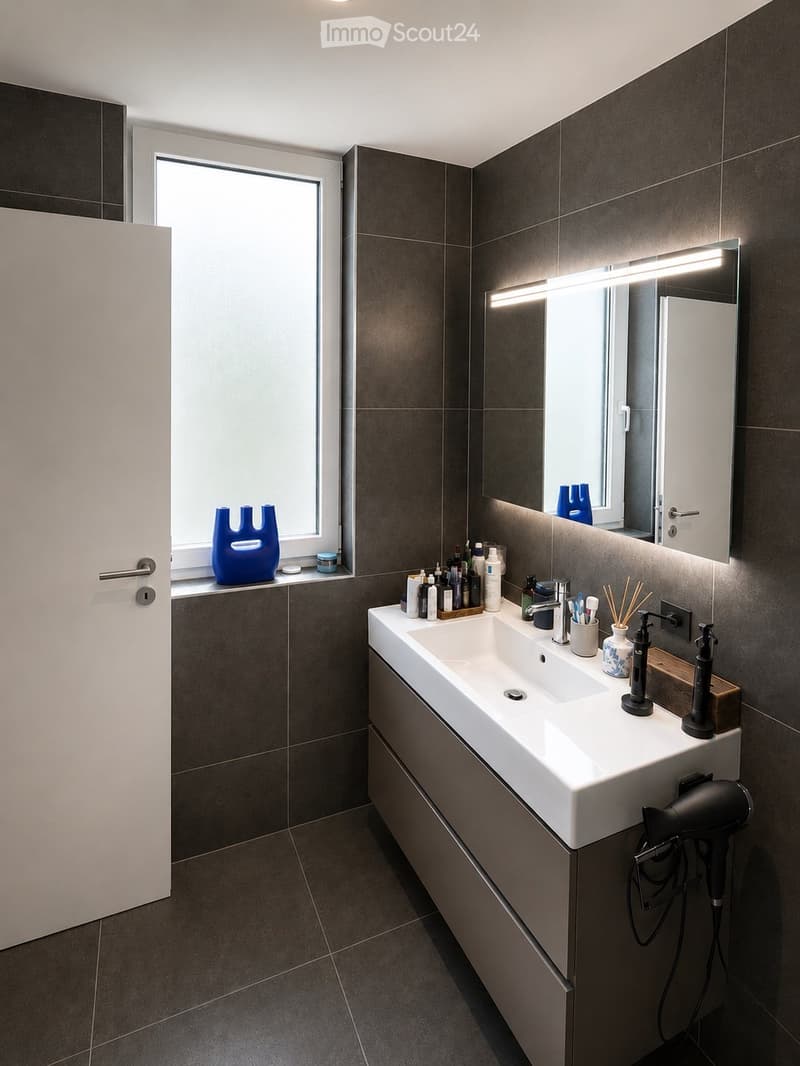 Modern bathroom with dark tiled walls and floor, white sink, large mirror with built-in lighting, and various toiletries on the counter.