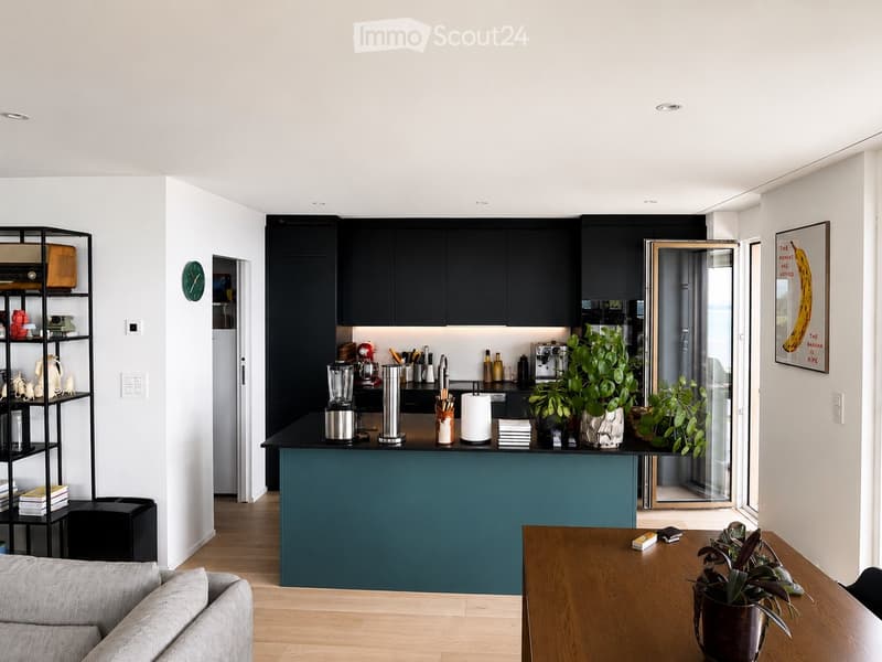 Modern open-plan living and kitchen area with a grey sofa, wooden coffee table, kitchen island, various appliances, and decorative items.