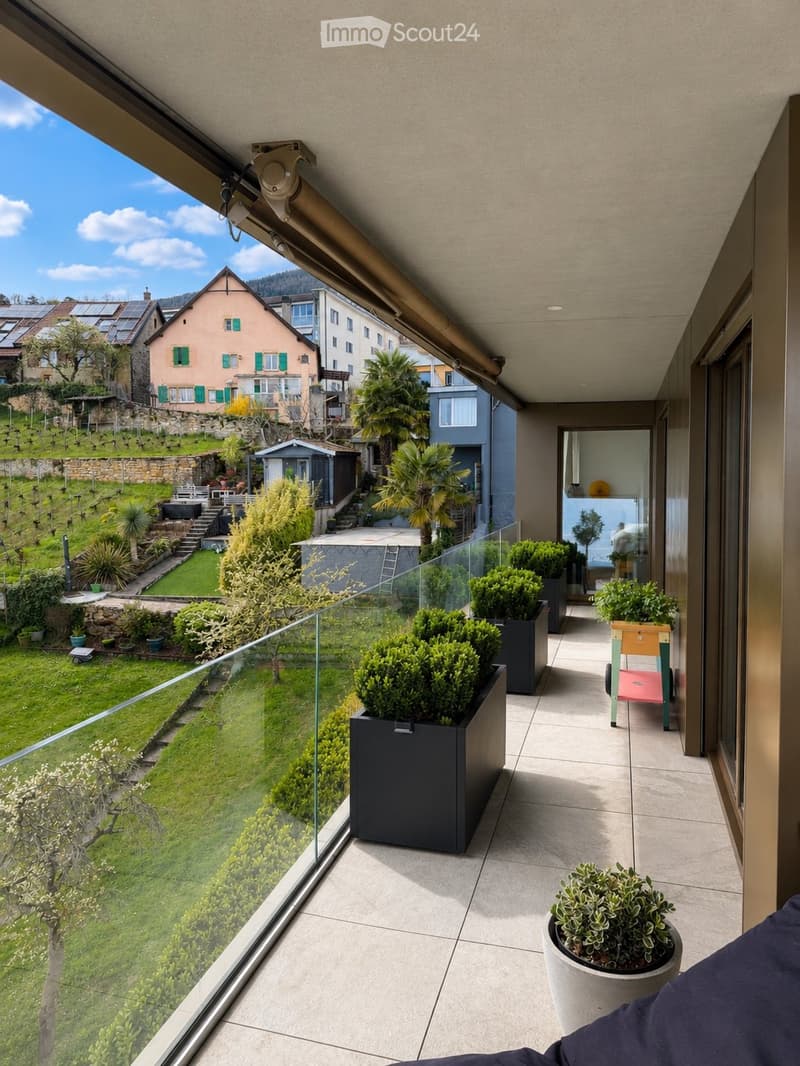 Modern balcony with tiled floor, glass railing, and potted plants, overlooking a landscaped garden and neighboring houses.