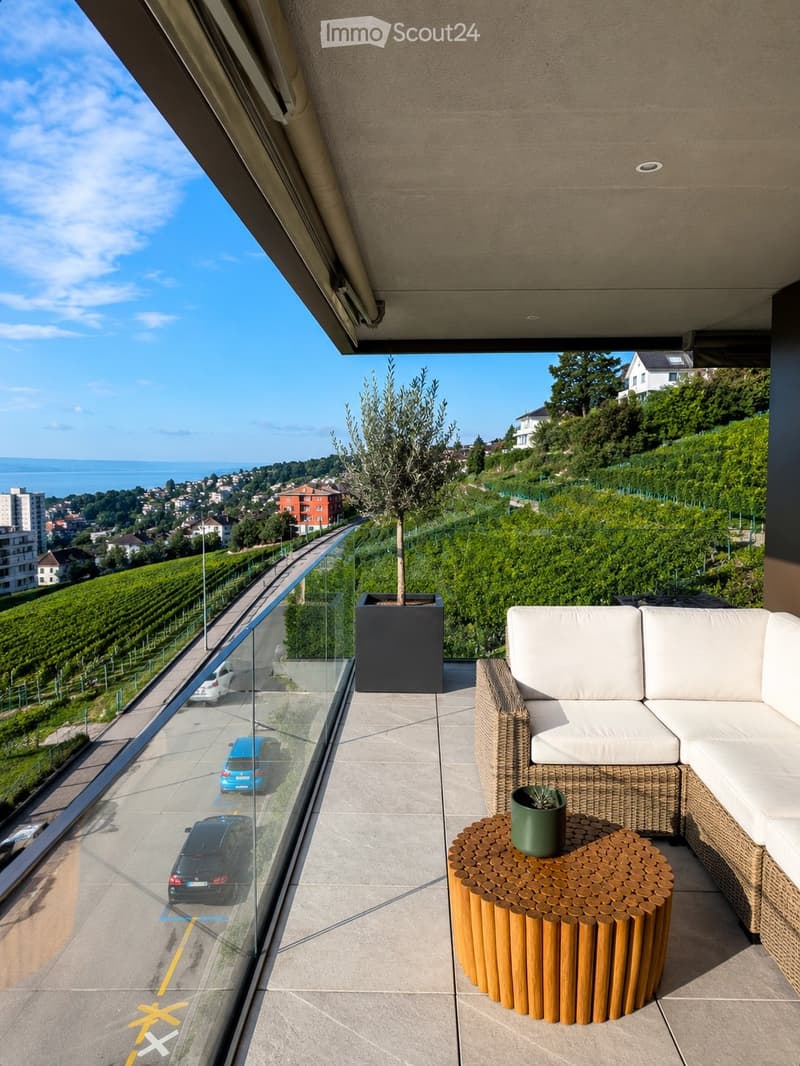 Modern balcony with a wicker sofa, round wooden table, and potted plants, offering a scenic view of the city and vineyards.