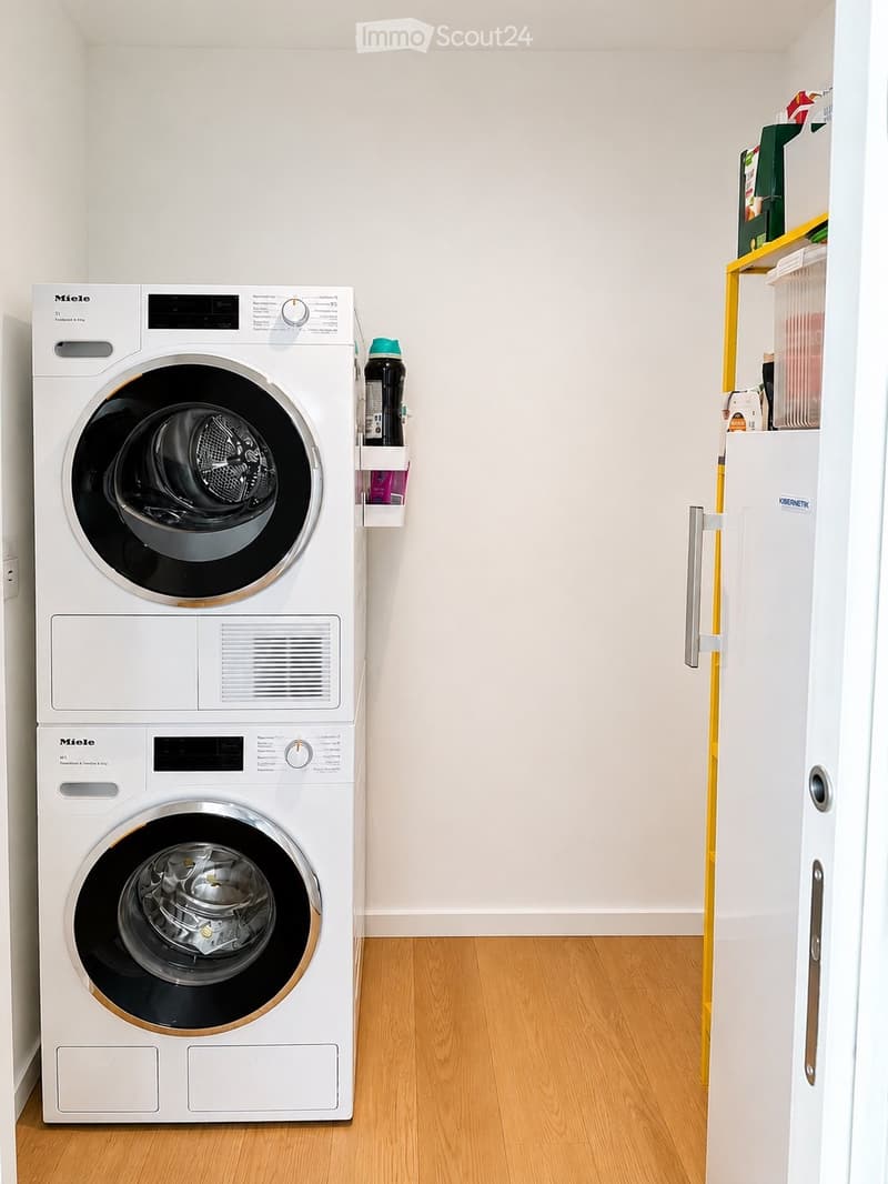 Bathroom with a washing machine and dryer stacked vertically, a small shelf with cleaning supplies, and a wooden floor.