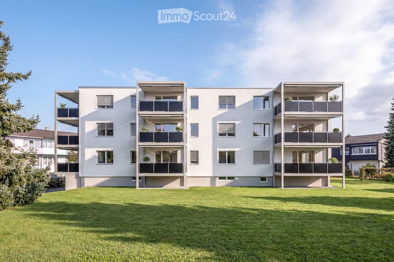 Modern multi-story apartment building with balconies and large windows, surrounded by a well-maintained lawn.