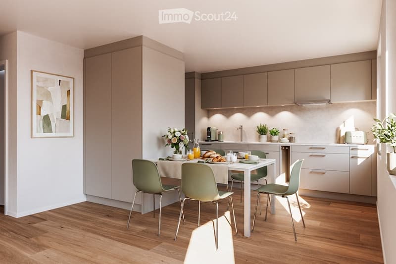A modern kitchen and dining area with light wooden flooring, white walls, and grey cabinets. A dining table with four green chairs is set with plates, glasses, and a vase of flowers. The kitchen features a sink, a refrigerator, and various small appliance