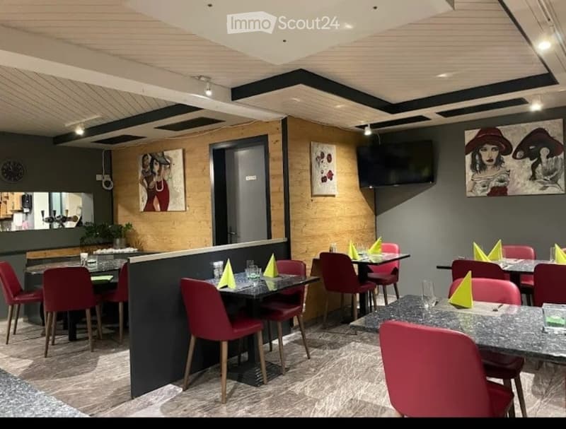Modern restaurant interior with dark grey walls, wooden accents, and red chairs around black tables. Green napkins, glassware, and a television mounted on the wall.