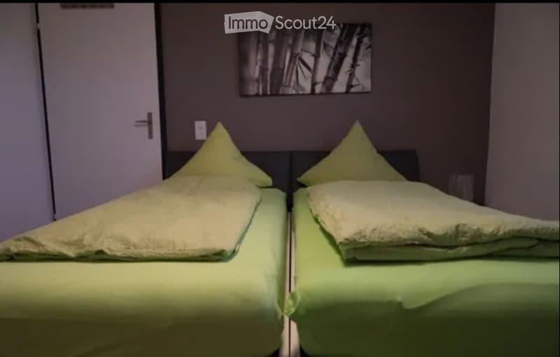 A small bedroom with two single beds, each with green bedding and yellow pillows, against a dark wall with a framed picture above.